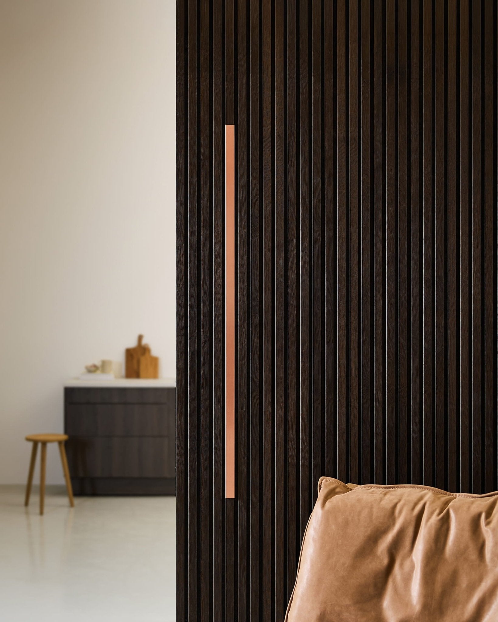 Modern room with a 120 cm Liva | Cover / Copper Akupanel acoustic wood slatwall by WoodUpp, vertical metal handle, tan chair, blurred bench and counter.