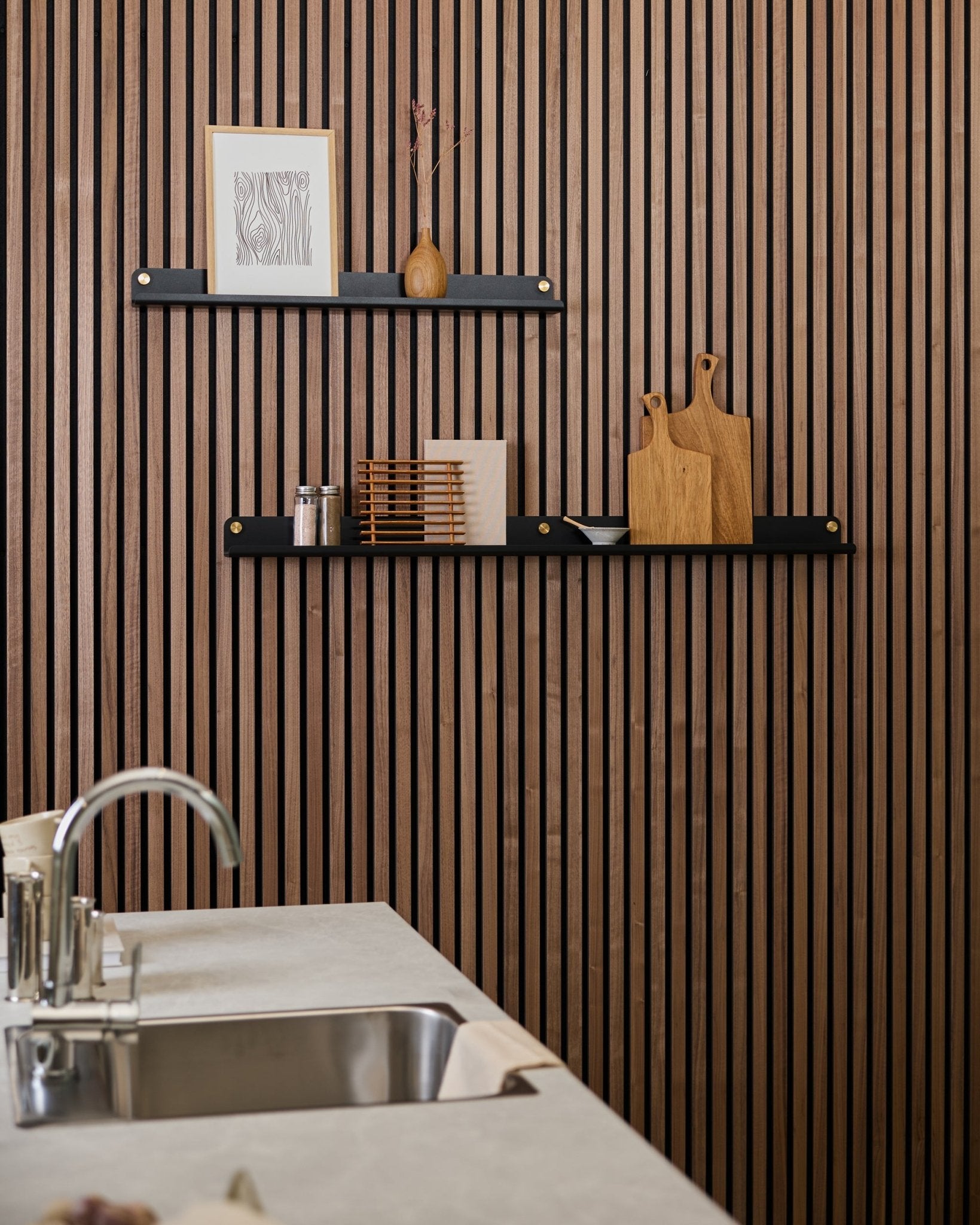 Minimalist kitchen with a vertical Akupanel acoustic wood slat wall, black shelves, cutting boards, small decor items, and a modern sink counter in front.