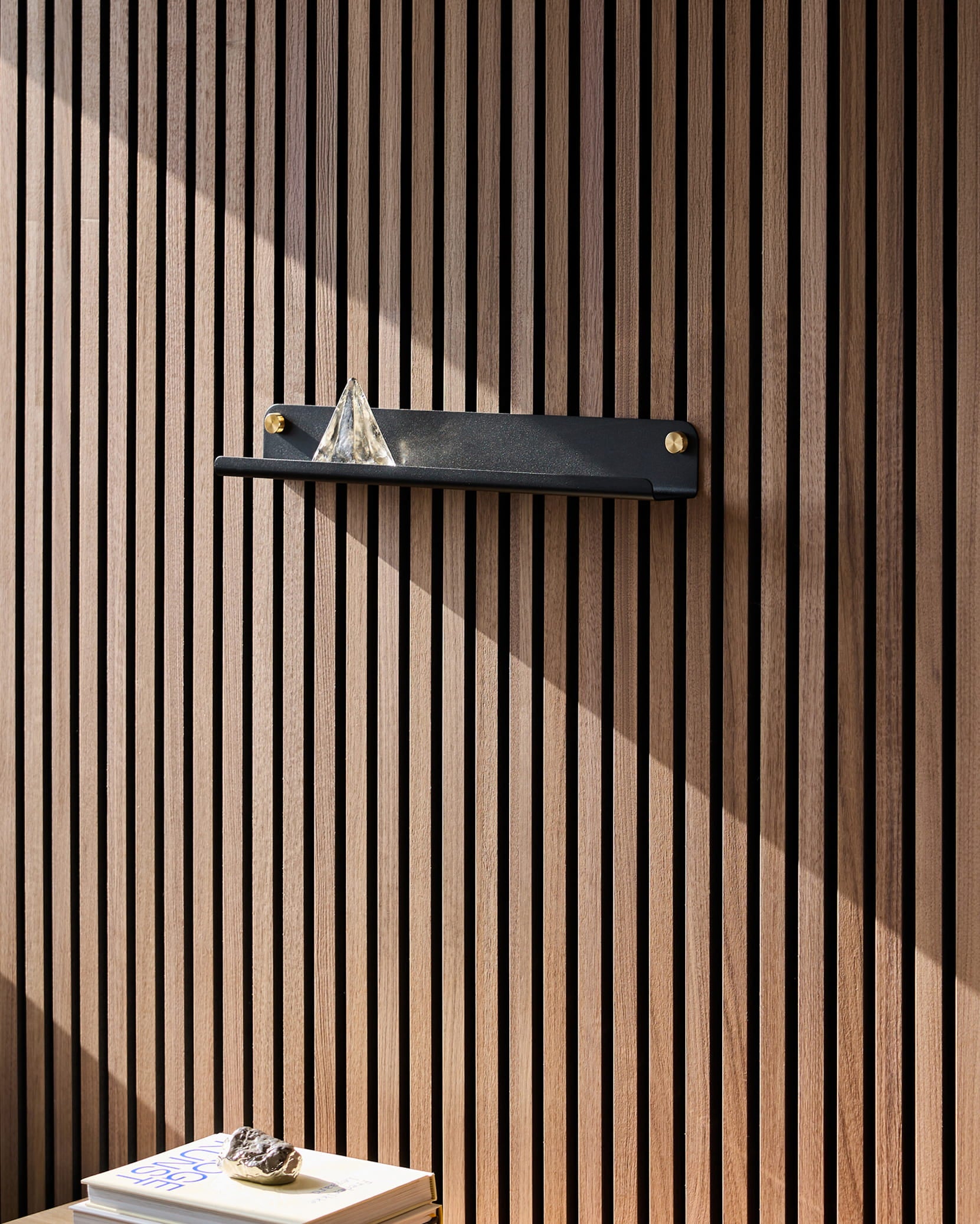 A black Norr Narrow Shelf by WoodUpp on an Akupanel acoustic wood panel holds a glass pyramid; sunlight hits books below, topped with a rock.