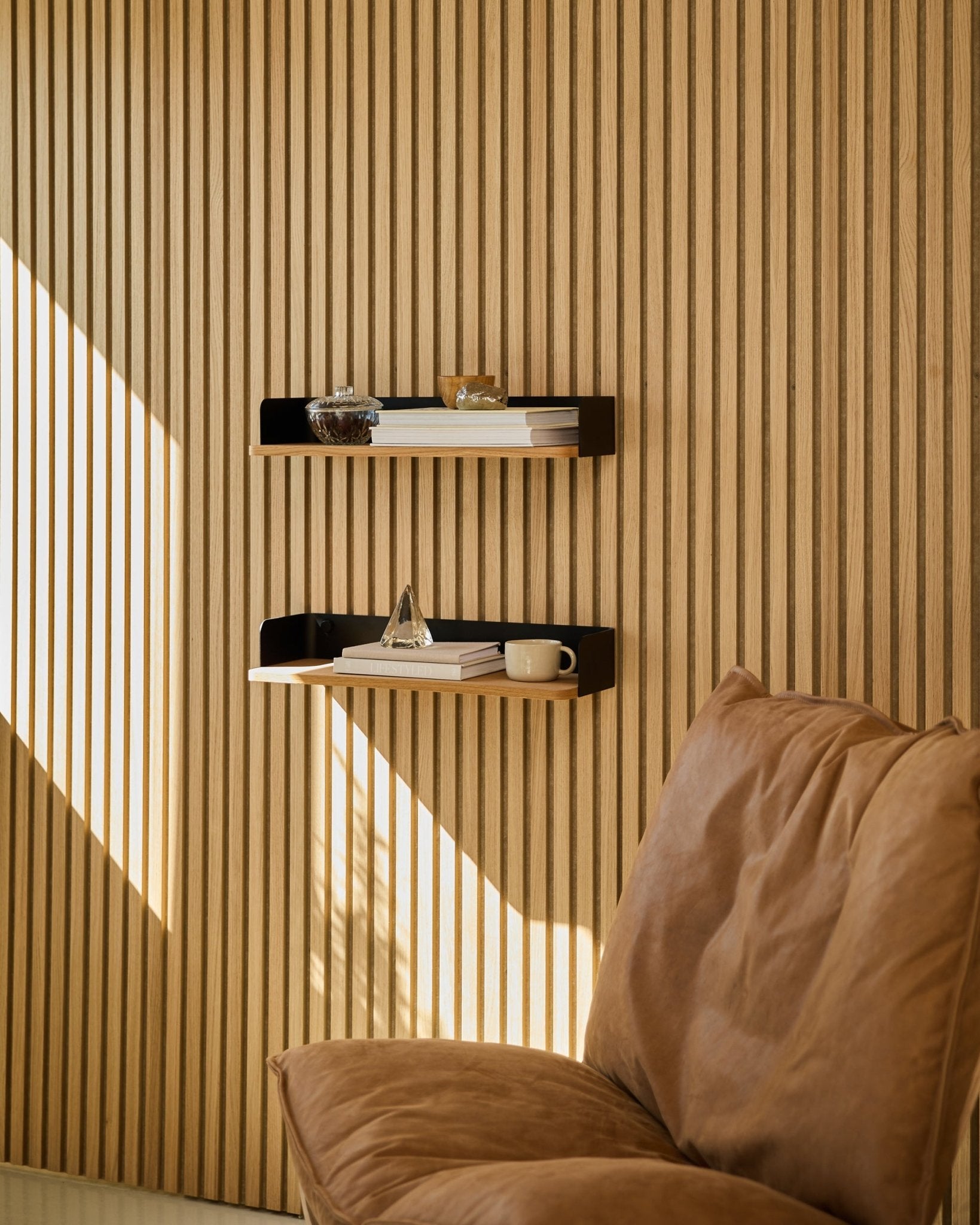 Brown chair next to the WoodUpp Linn Bookshelf in Natural Oak, with books and decor arranged in natural light by a Nordic wood slat acoustic panel wall.
