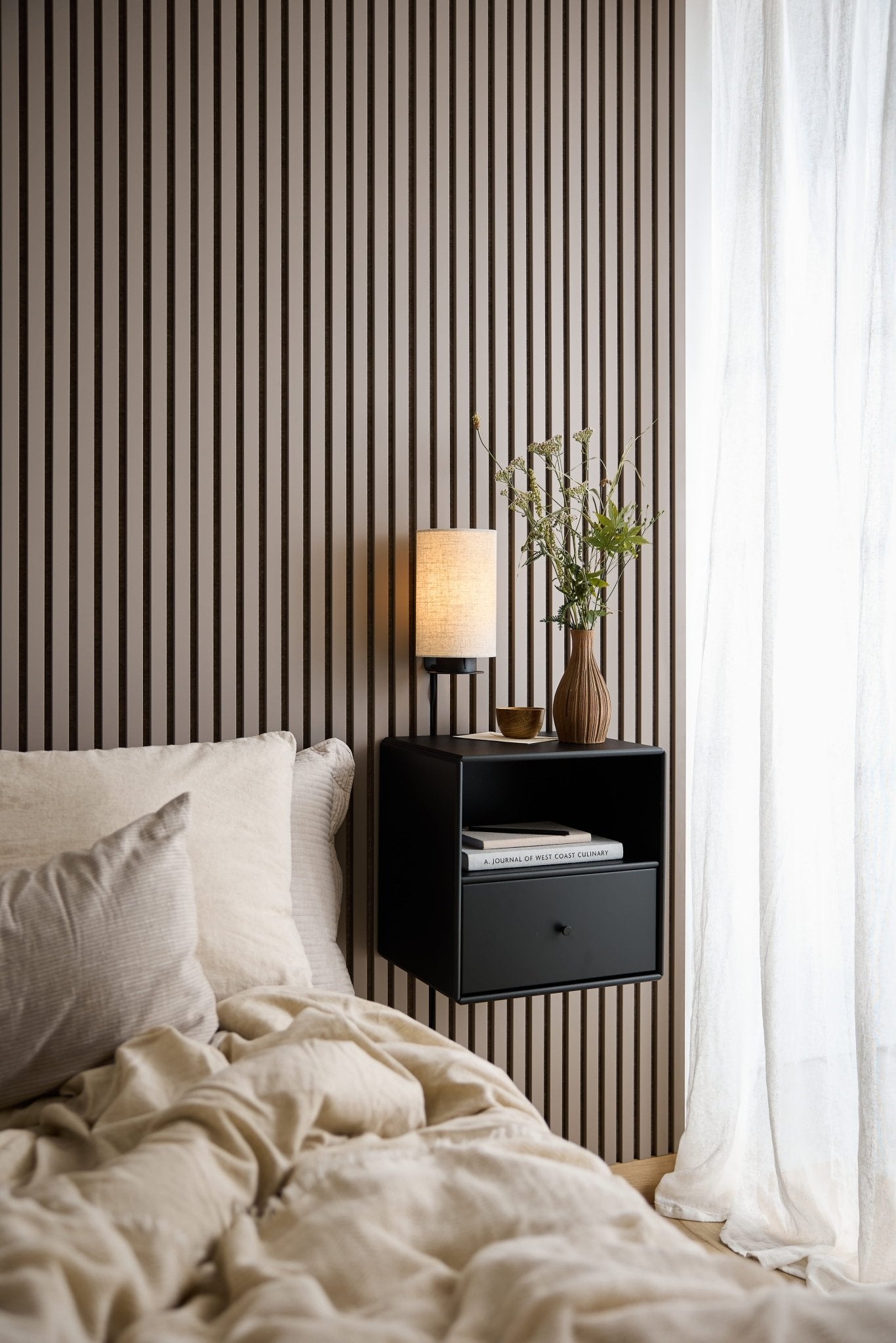 Bed with beige bedding, black WoodUpp Arka Bookcase Drawer, lamp, and vase near a window with sheer white curtains.