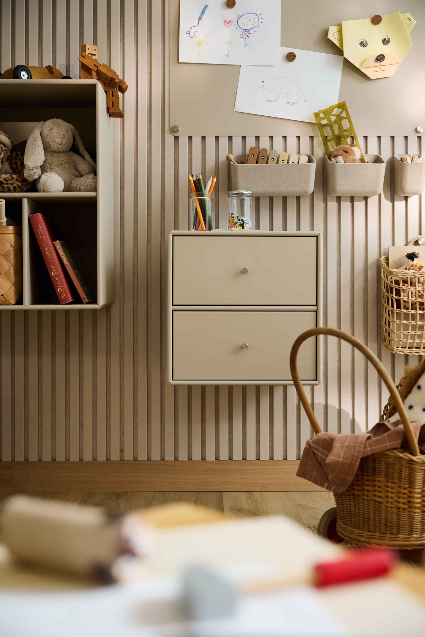 Children’s room with striped wallpaper, toys, books, baskets, and a beige WoodUpp Arka Bookcase Drawer providing closed storage.