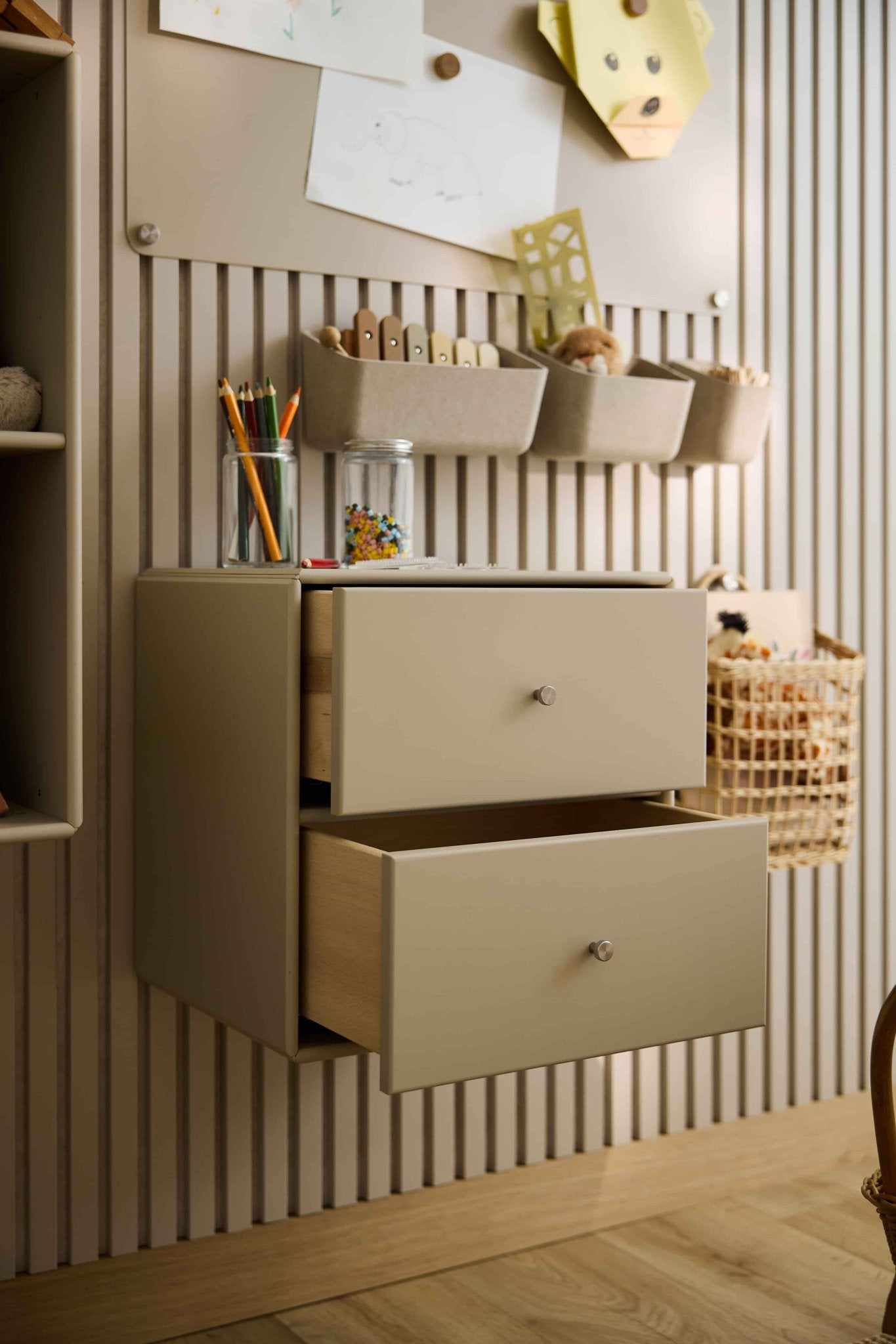 Two beige Arka Bookcase Drawers by WoodUpp, partially open, store art supplies and toys; closed storage and baskets are placed nearby.