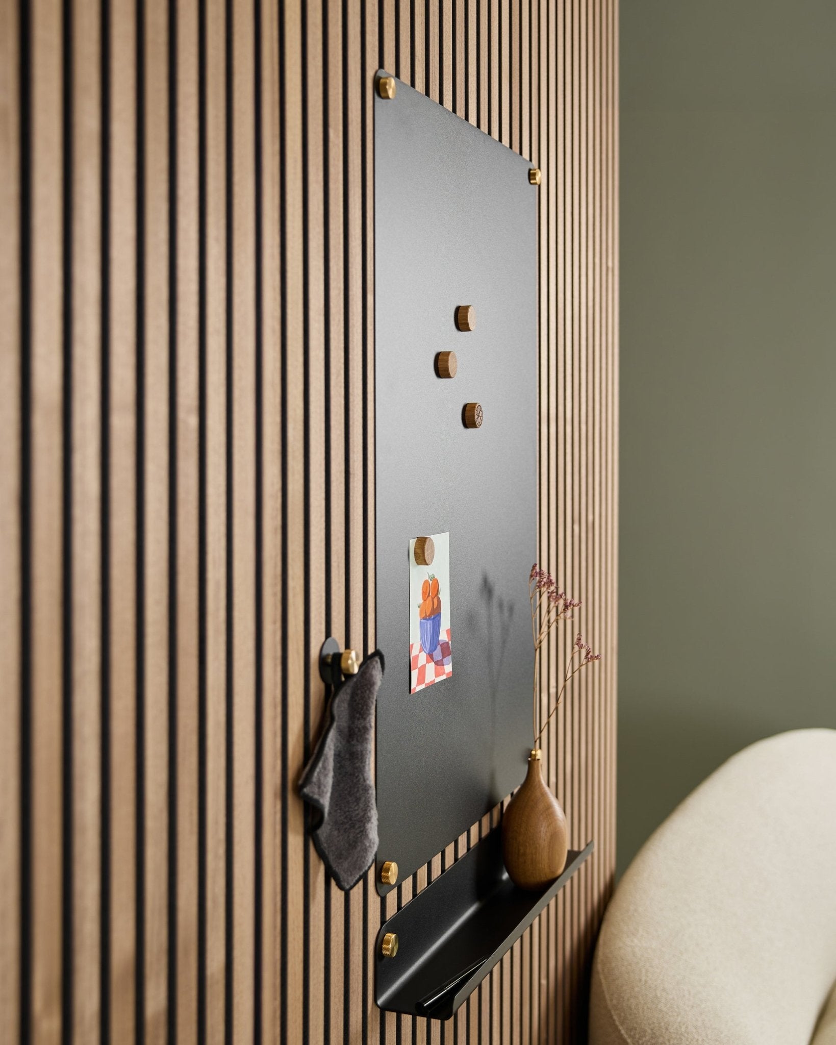The WoodUpp Norr large black acoustic magnetic board with wooden magnets, pinboard, shelf, and vase is mounted on the wall beside a beige chair.