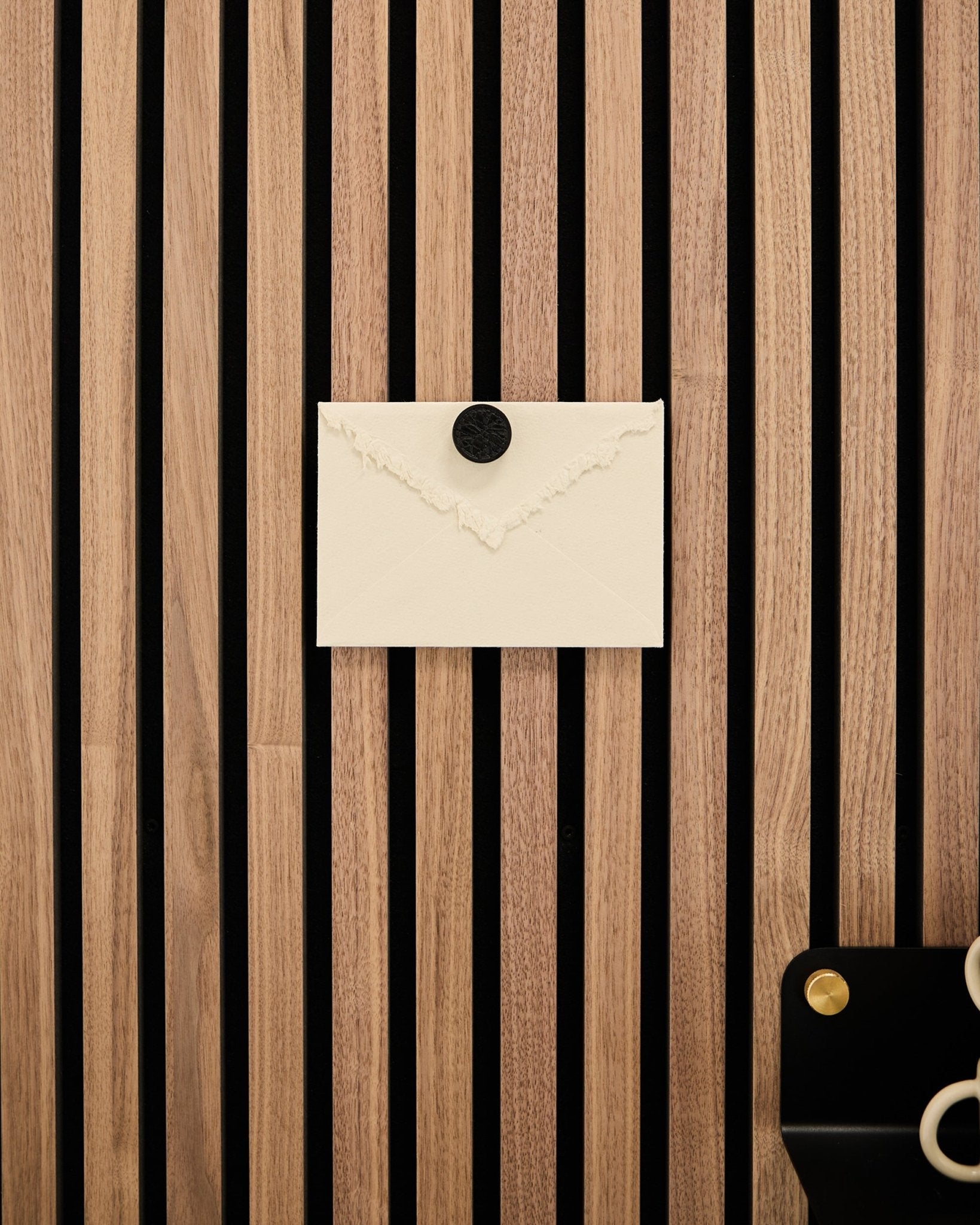 A cream envelope is pinned to a WoodUpp Akupanel oak-striped acoustic panel using the Norr Poster Magnet set in Black Oak; scissors rest at the bottom right.
