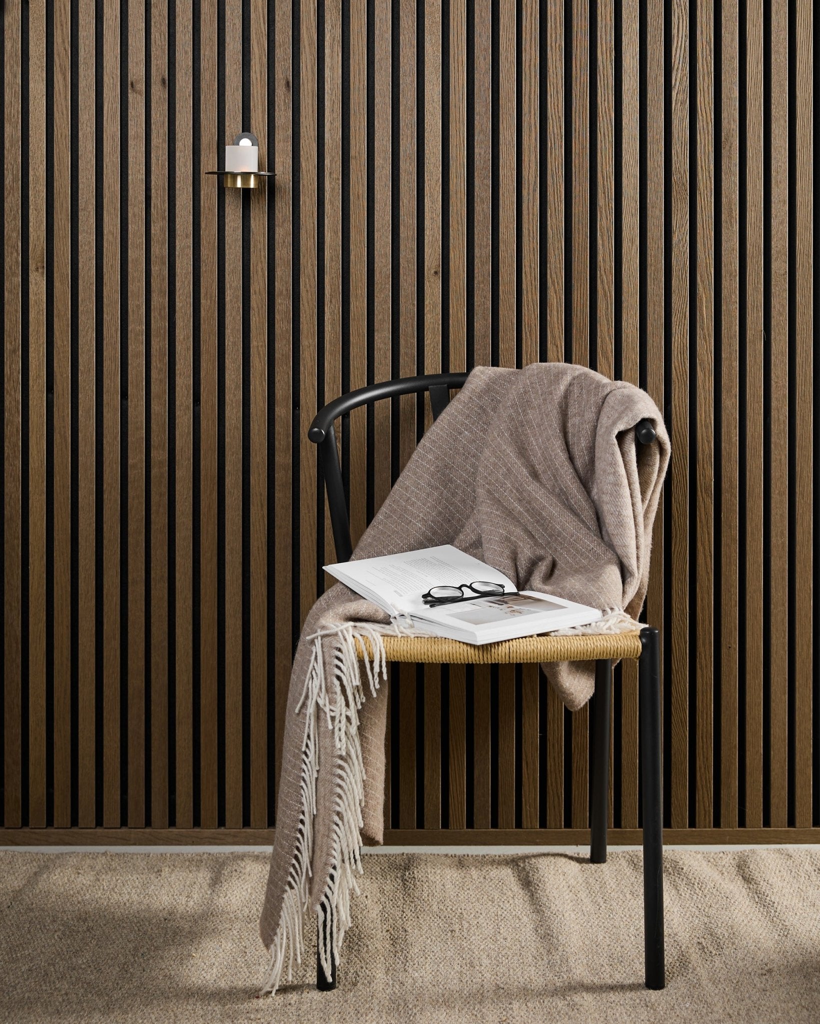 A modern chair with a beige throw blanket, open book, and eyeglasses sits by an Akupanel acoustic wood panel and a black and brass WoodUpp Norr Candle Holder.