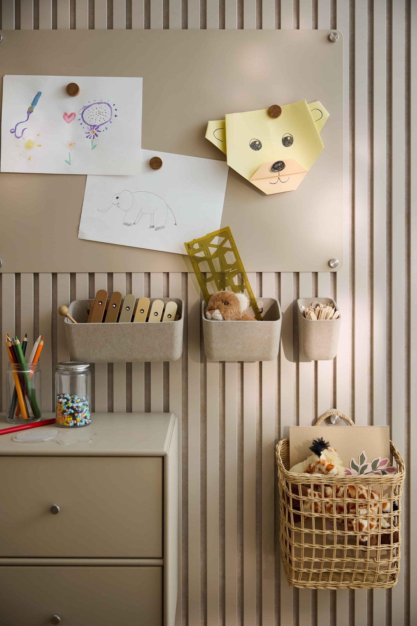 Children’s art and toys are arranged on a beige wall with WoodUpp acoustic panel, featuring felt containers, baskets, a drawer, and a pegboard.