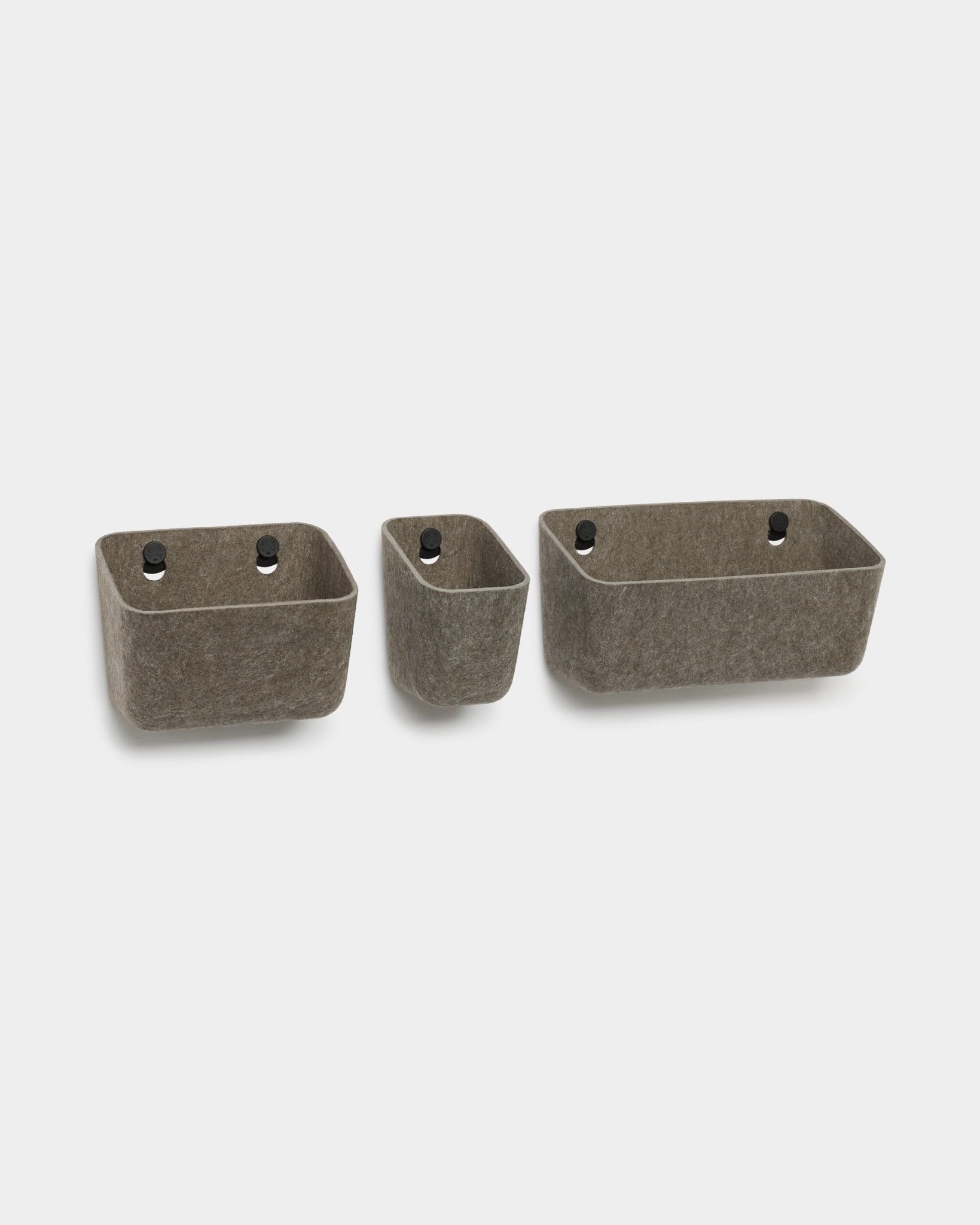 Tre contenitori in feltro WoodUpp Luva color grigio scuro caldo con occhielli neri, progettati per essere montati a parete come organizer compatibili con i pannelli acustici WoodUpp.