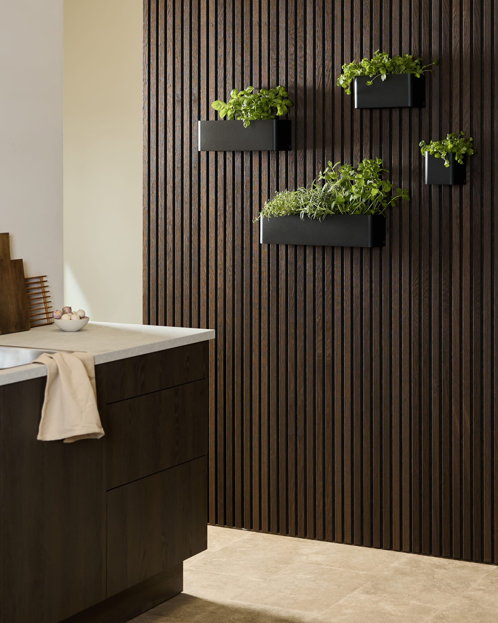 Modern kitchen featuring a WoodUpp Akupanel dark wood acoustic panel, black Norr Plant Box with herbs, and dark cabinetry topped with a white countertop.