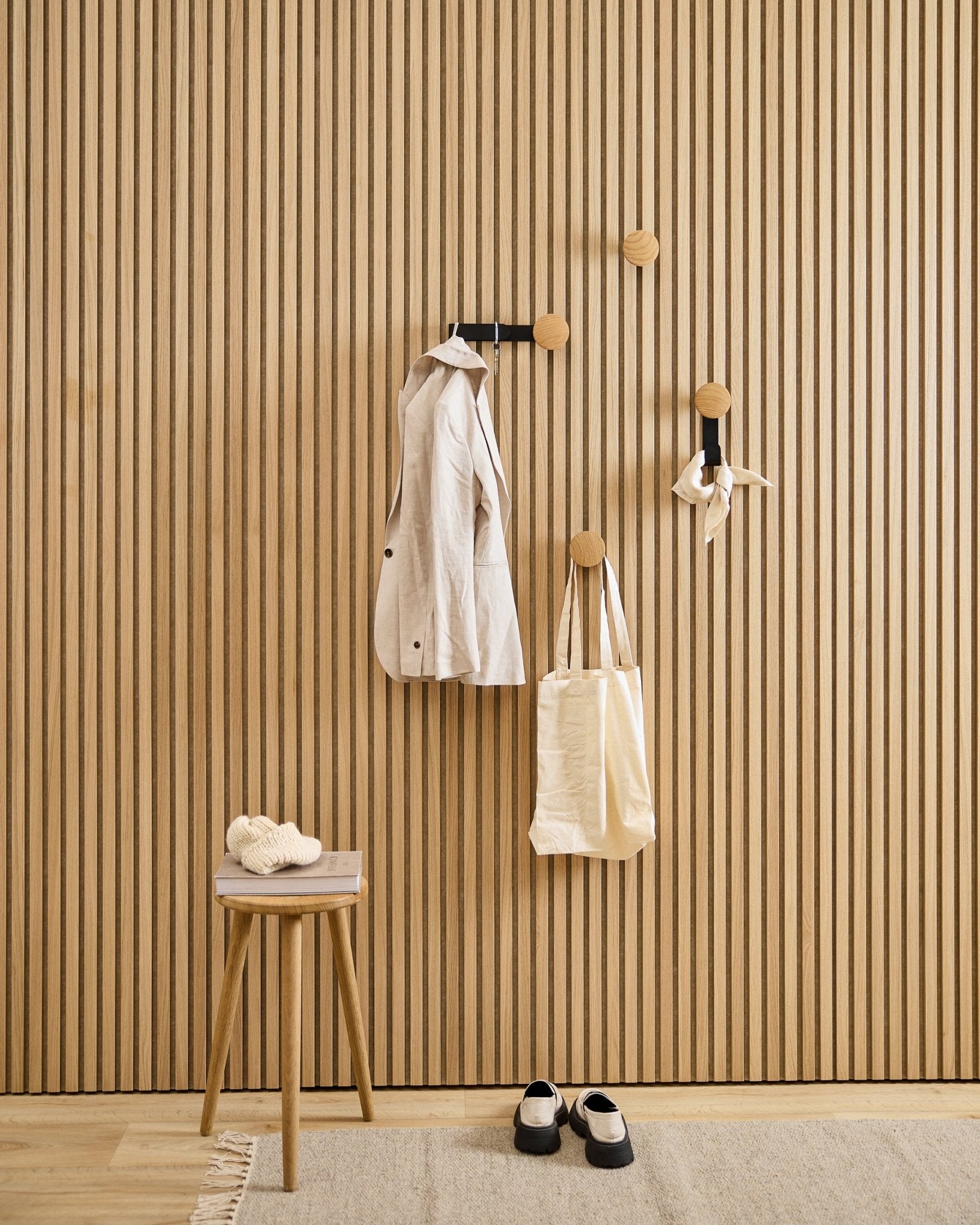 Minimalist entryway featuring WoodUpp Linn Vertical Hanger in natural oak, with a beige coat, tote bag, scarf, stool holding slippers, and black-and-white shoes.