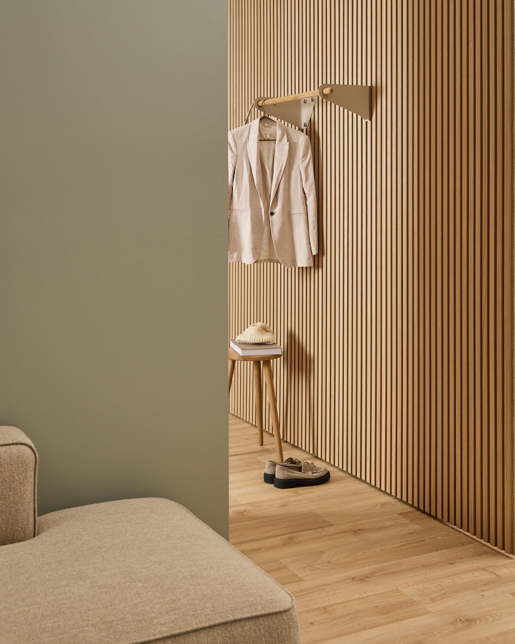 Minimalist room with beige sofa, Akupanel acoustic slat wall, WoodUpp Linn Coat Rail in beige (75.9 cm), stool with hat, and shoes on light wood floor.