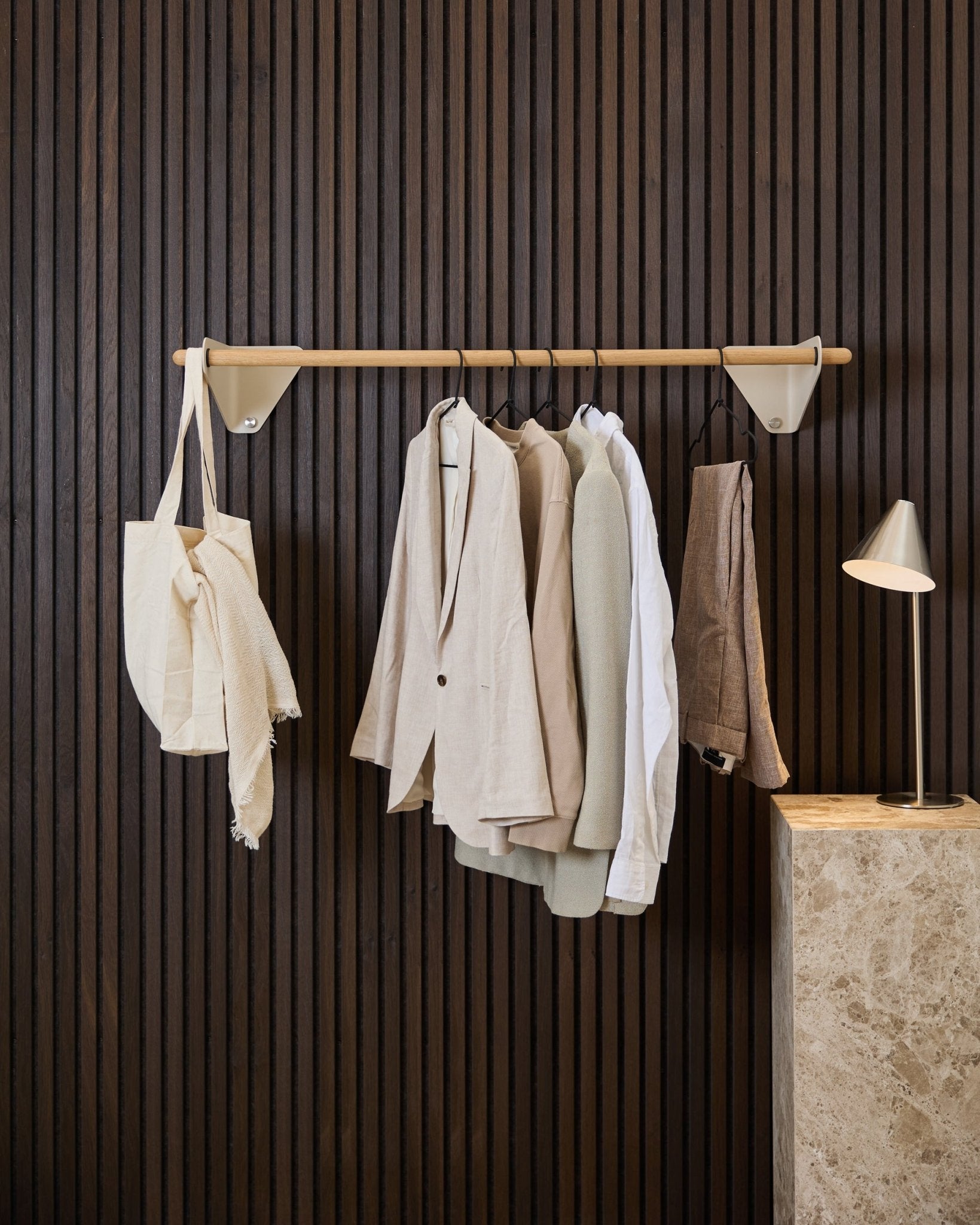 A beige WoodUpp Linn Coat Rail (115.9 cm) holds jackets, a white shirt, and a cream tote in front of a dark wood Akupanel acoustic panel and marble lamp.