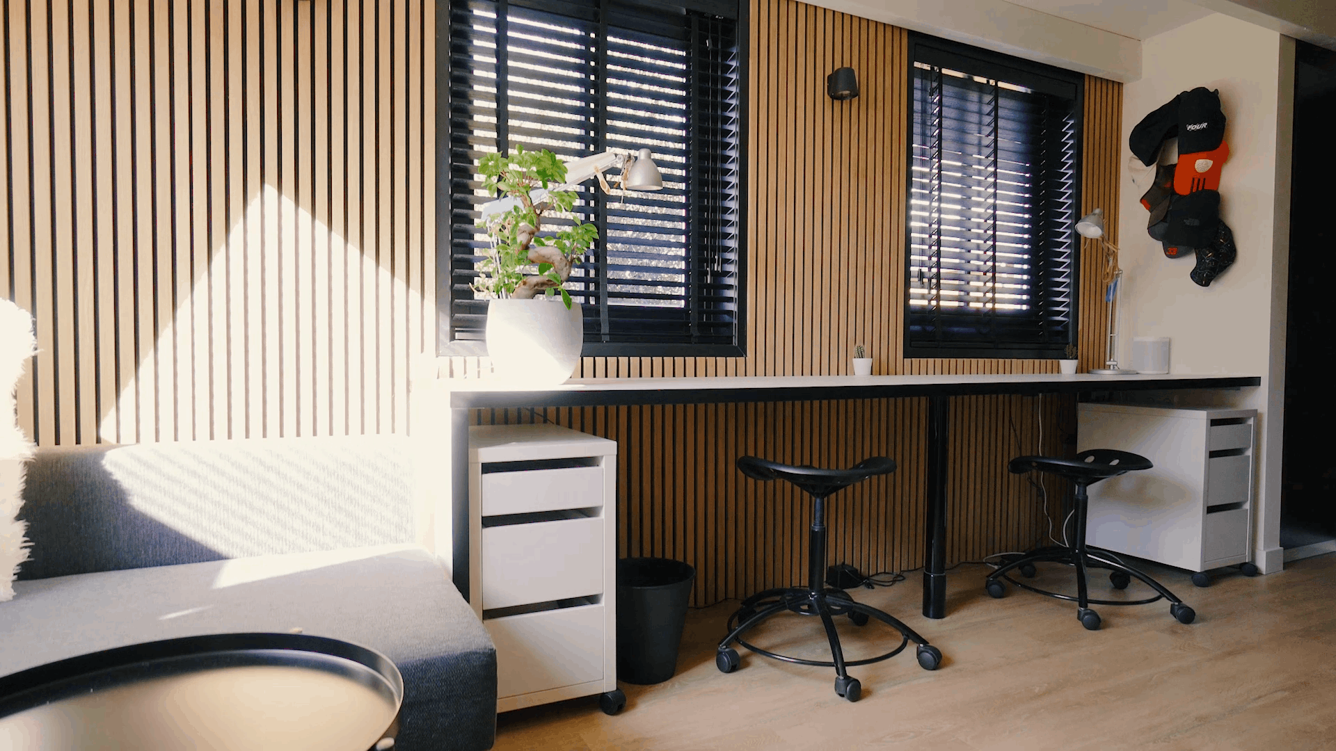 Modern office featuring Nordic acoustic wood panels on walls, ergonomic workstations, black stools, a potted plant, and sunlight through black window blinds.