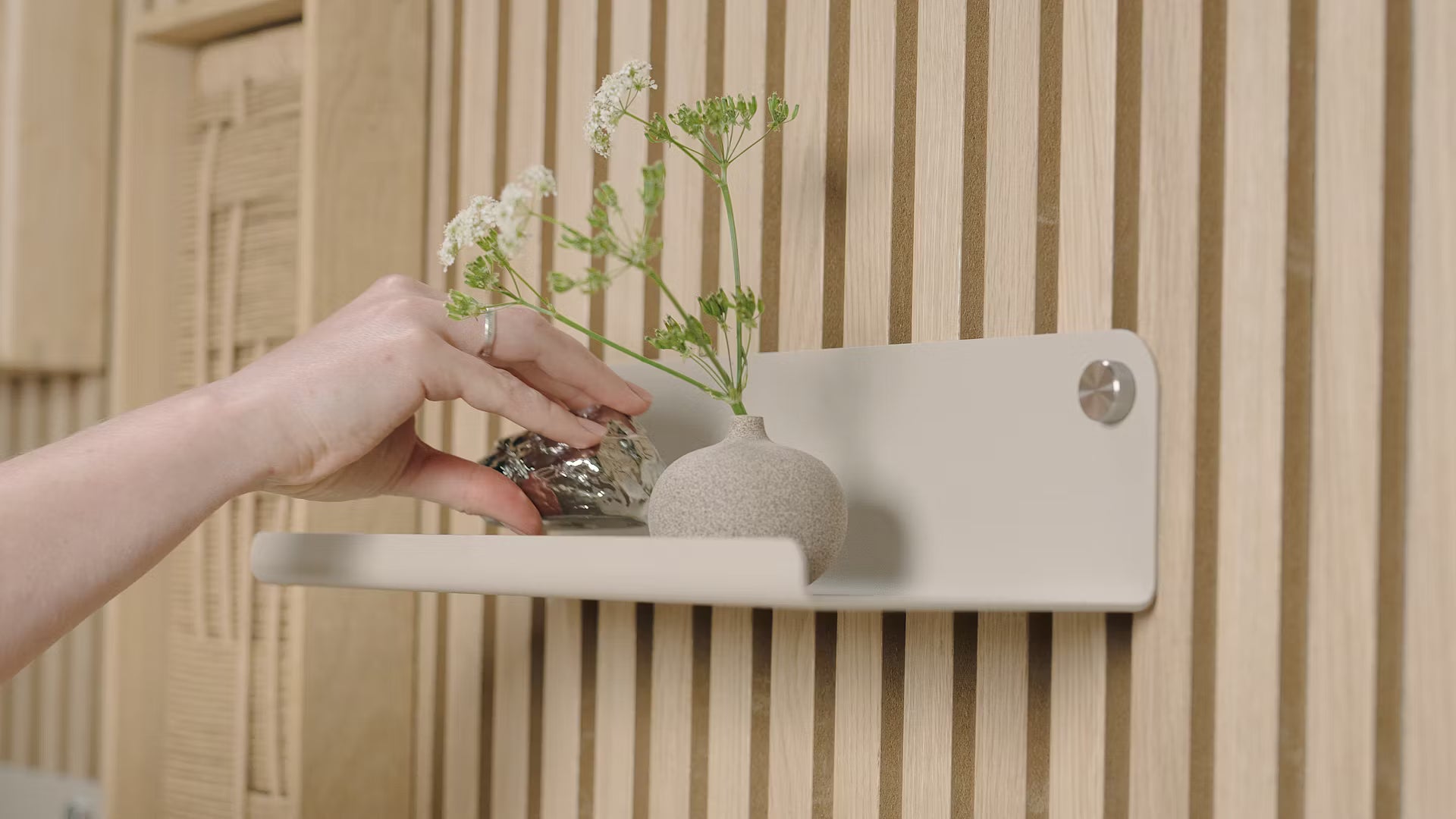 A hand places a small decorative object beside a vase of white flowers on a minimalist shelf set against a textured Nordic acoustic wood slatwall panel.