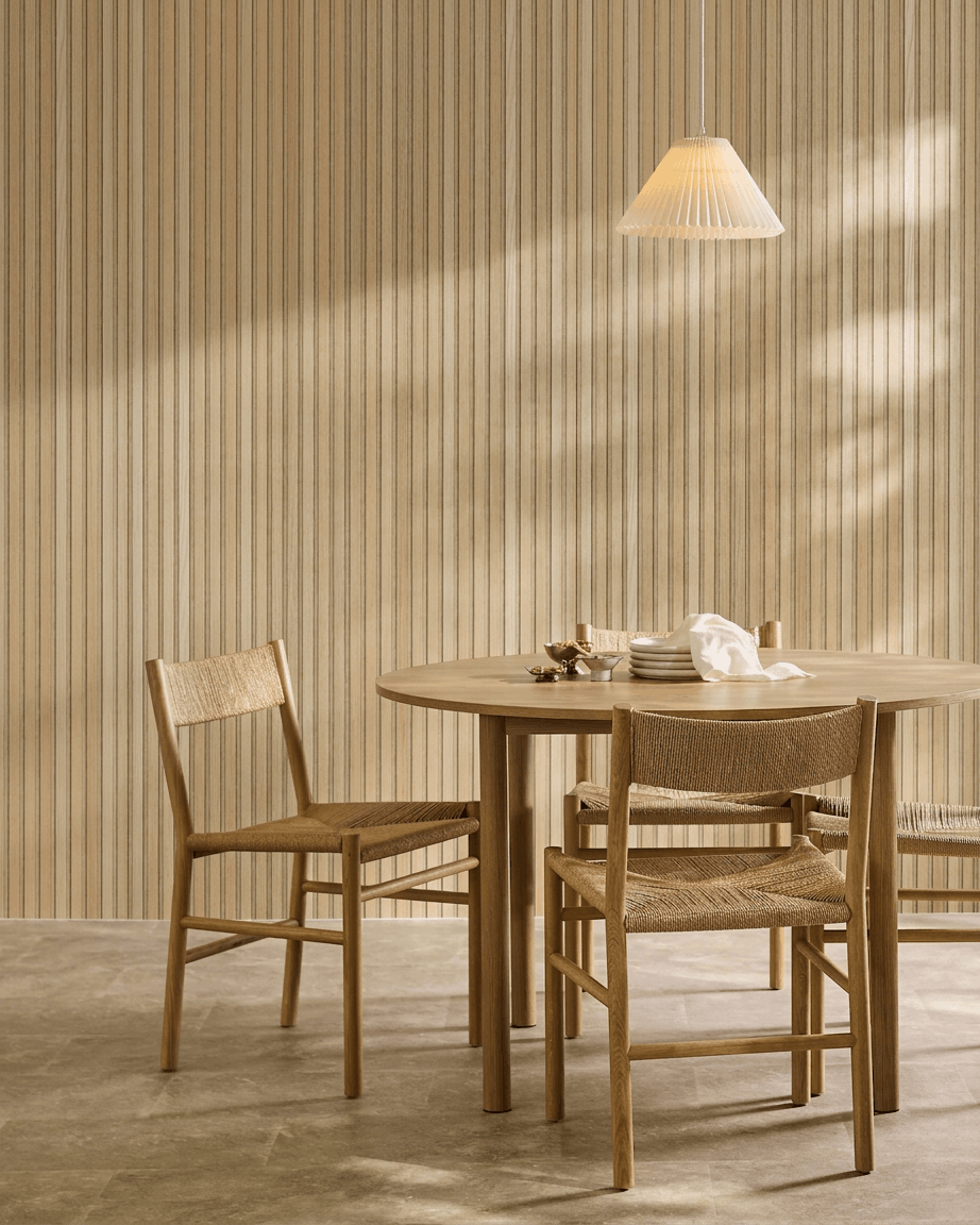 Minimalist dining area with a round table, woven chairs, and pendant light beside a WoodUpp Akupanel acoustic wood panel in Classic Oak with beige felt.