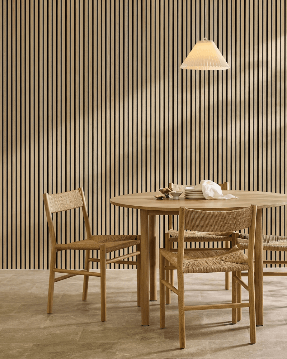 Minimalist dining area featuring a round table, woven chairs, pendant lamp, and a WoodUpp Akupanel Classic Oak acoustic panel mounted on the wall.