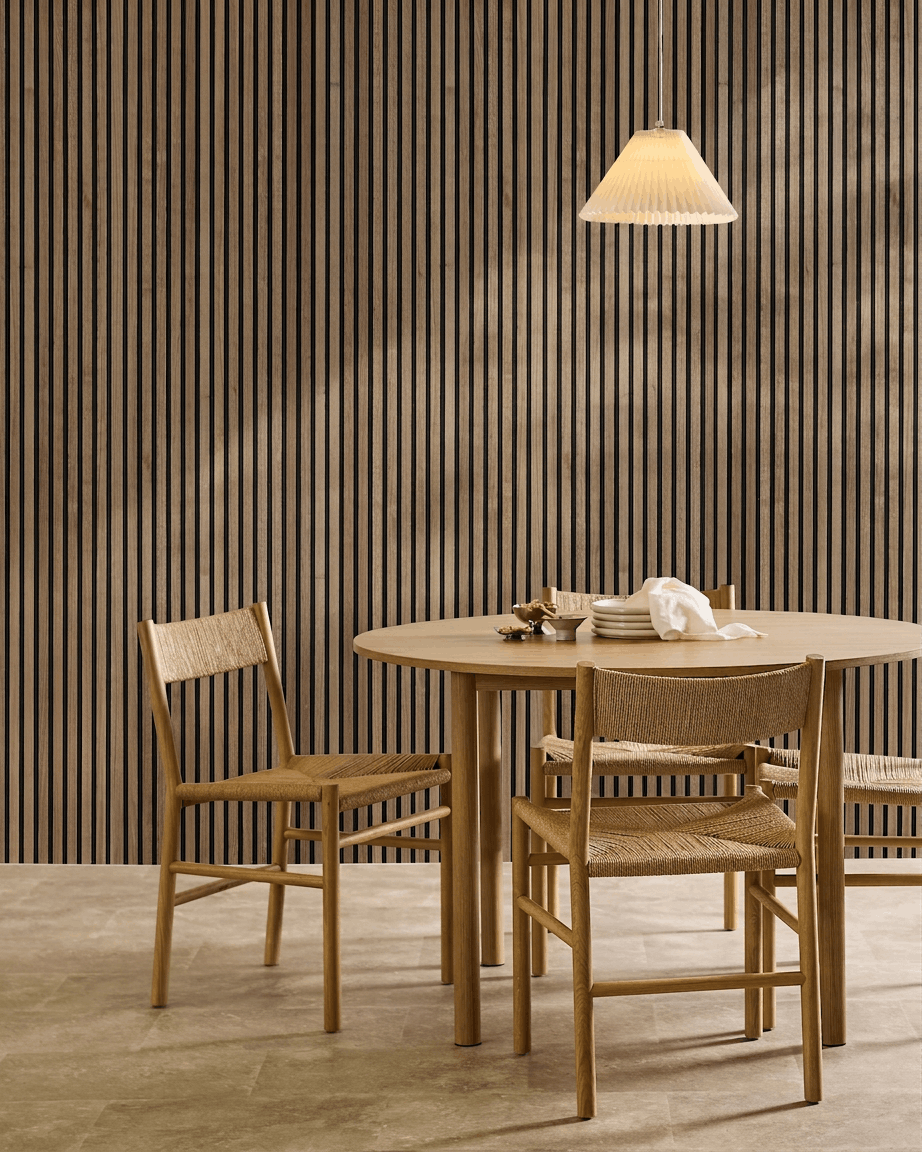 Minimalist dining area with a round table, woven chairs, pendant lamp, and a WoodUpp Akupanel acoustic wood panel in Natural Wood with black felt on the wall.
