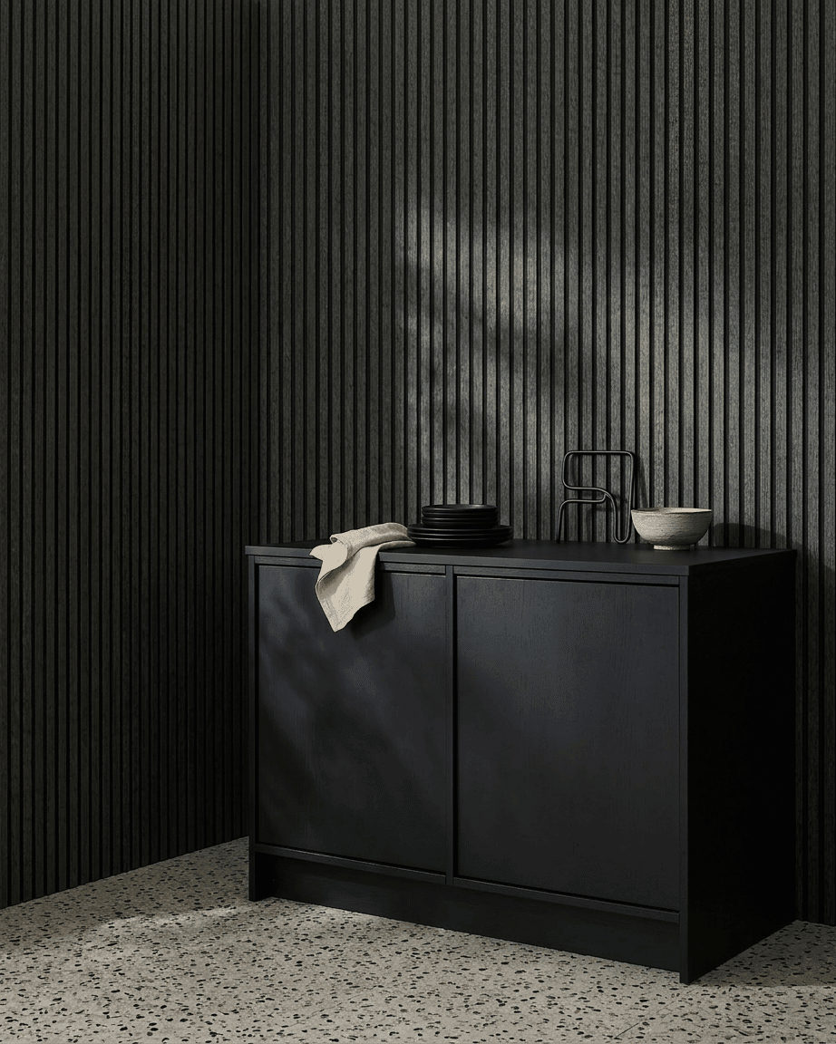 A black cabinet next to a WoodUpp Akupanel acoustic wood panel in Charcoal/Black Felt, topped with bowls and plates, on a light terrazzo floor.