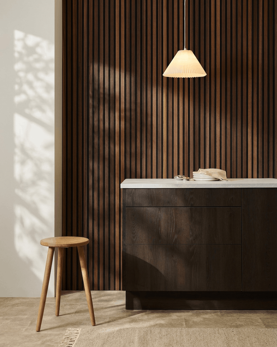 Minimalist interior featuring a wooden stool, dark wood cabinet, pendant lamp, and a WoodUpp Akupanel acoustic wood slatwall panel in Natural Wood/Smoked Larch.