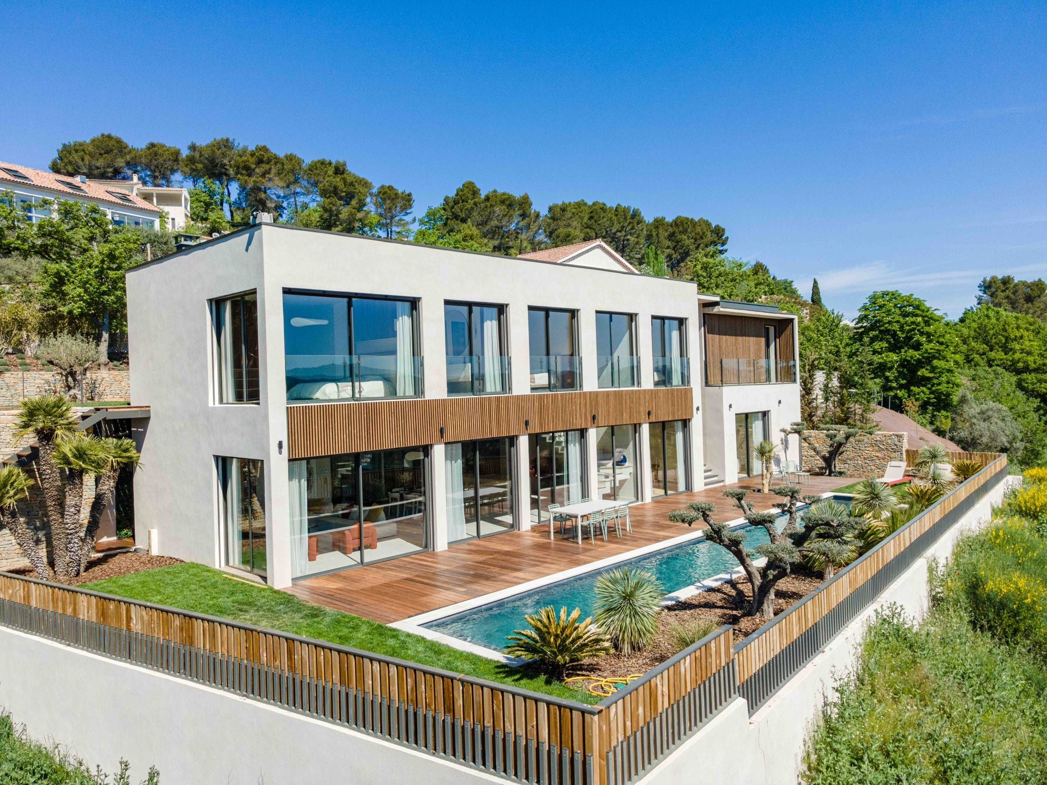 Modern two-story house with large windows, Nordic wooden facade panels, a pool, spacious deck, and greenery under a clear blue sky.