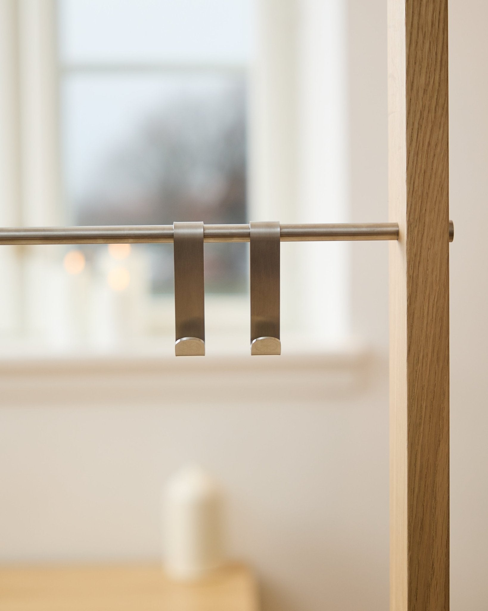 WoodUpp Alva S-Hook BM1  BM2 (2 pcs) in stainless steel, hanging on an acoustic wood panel rod; blurred window and lit candles in the background.
