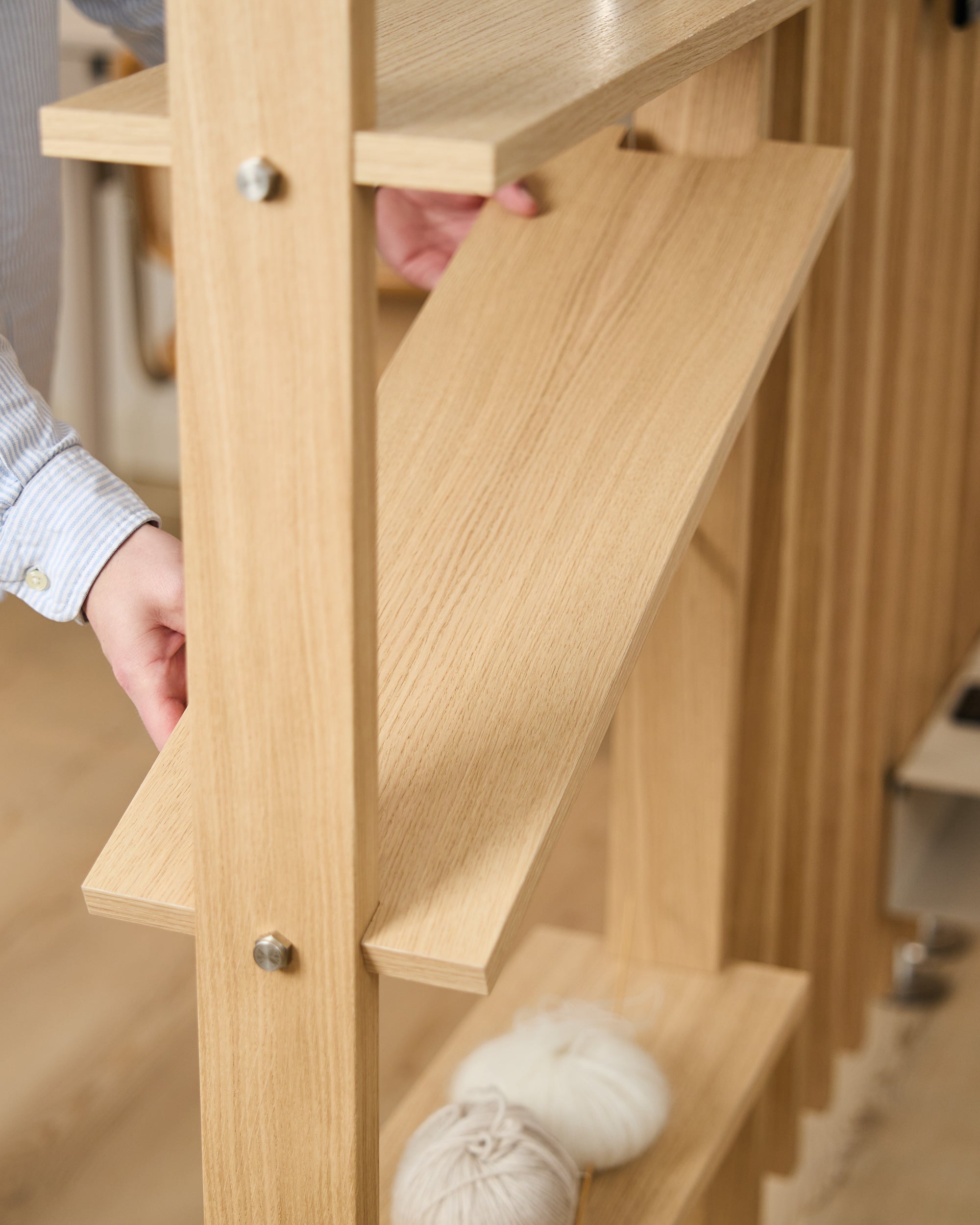 CREATE wooden shelf accessory being installed on a light wood shelving frame, yarn balls on the lower shelf