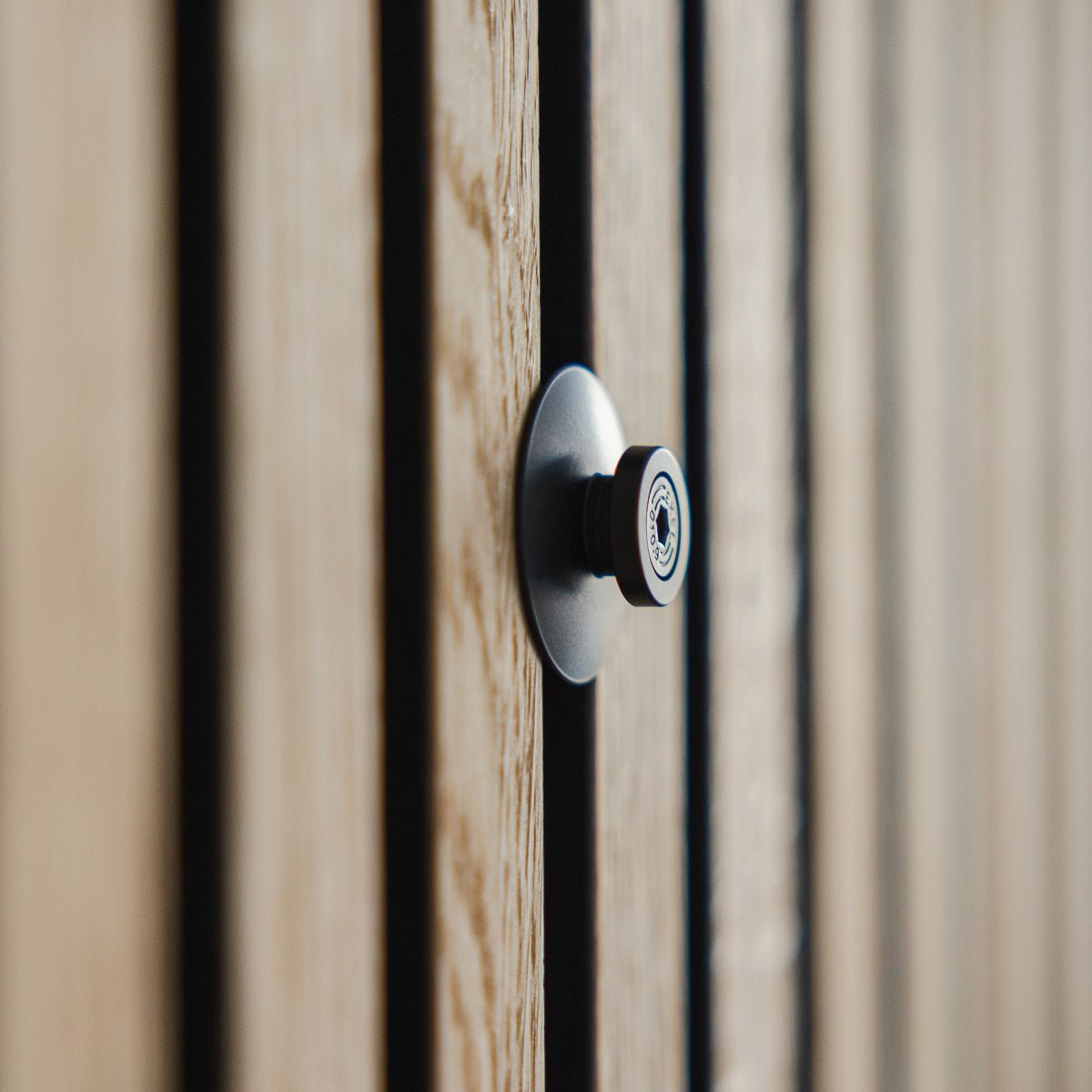 Close-up of the Dupps | Heavy peephole by WoodUpp in black aluminium, installed on a grooved wooden door with Akupanel acoustic slats, lens in focus.