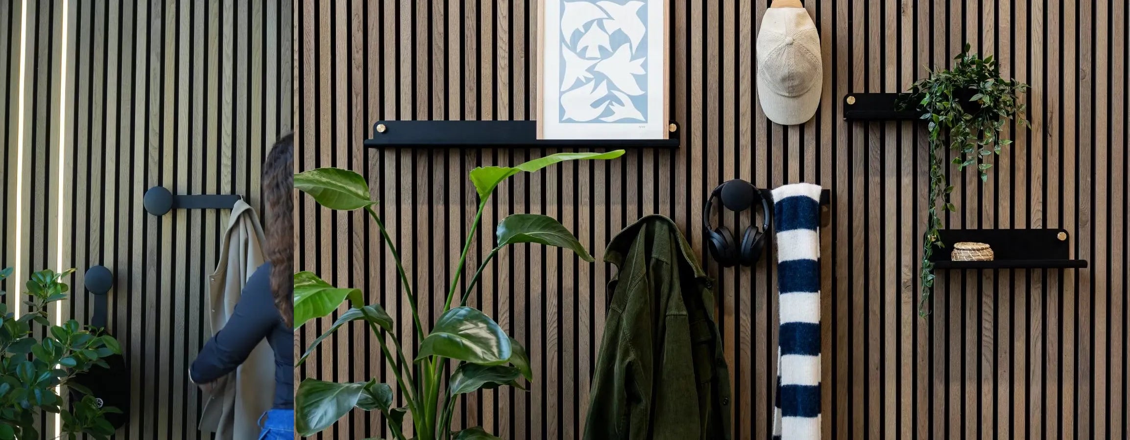 A man in a blue jacket walks past a Nordic Akupanel acoustic wood slatwall with integrated plants, shelves, coat hooks, headphones, and a rolled striped towel.